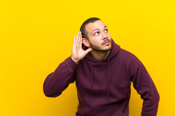 Colombian man with sweatshirt over yellow wall listening to something by putting hand on the ear