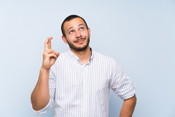 Colombian man over isolated blue wall with fingers crossing and wishing the best