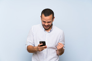 Colombian man over isolated blue wall with phone in victory position