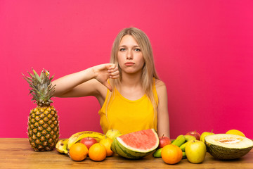 Young blonde woman with lots of fruits showing thumb down sign