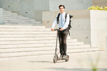 Young businessman with backpack on his back riding on scooter to work along the street in the city