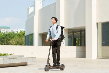 Young smiling manager with backpack behind his back riding on electric scooter past the office building outdoors © DragonImages