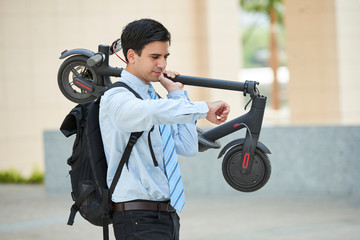 Young businessman standing with backpack behind his back holding electric scooter and looking at the time on watch on his hand while standing in the city. © DragonImages