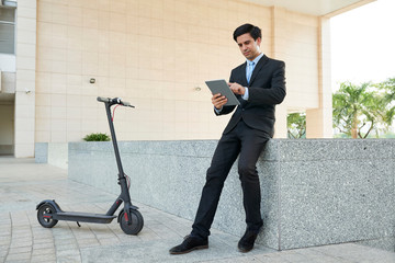 Young businessman in suit working online on digital tablet while sitting in the city with electric...