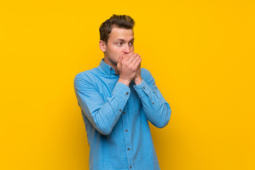 Blonde man over isolated yellow wall covering mouth and looking to the side