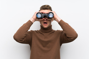 Blonde man over isolated white wall and looking in the distance with binoculars