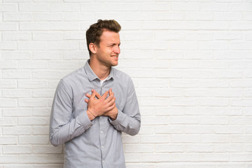 Blonde man over white brick wall having a pain in the heart