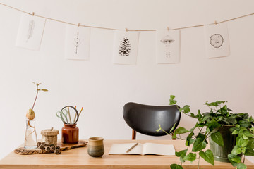 Scandinavian interior of home office space with wooden desk, design chair, forest accessories, avocado plant, office supplies. Stylish drawings on the white wall. Botanical home decor. Minimalistic.