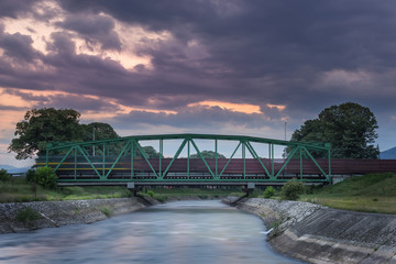 Fototapeta premium Blurred motion, long exposure of train passing over metallic railway bridge during blue hour with sky still dramatic after sunset