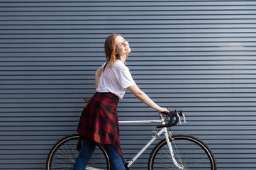 beautiful young girl standing with a white bicycle on a background of gray striped wall, the woman is happy