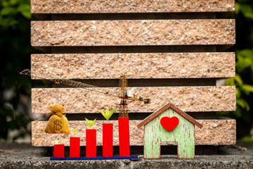 Fototapeta premium Home model with red heart and bar graph with growing value and tower crane with money bag put on the on the wall in the public park, Saving money and loan for construction real estate concept.