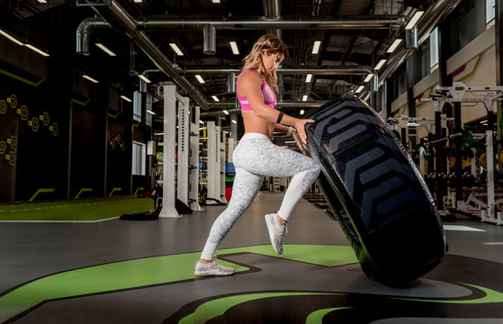 Beautiful Blonde fitness athlete wearing long leggings and pink sports bra flipping a large tire in a gym