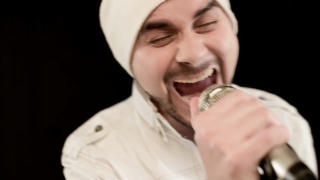 Close-up Frontman vocalist rock pop with a stylish beard in white clothes and a hat with a microphone in his hands expressively aggressively singing in the studio against the background of black walls