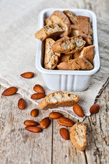 Cookies cantuccini in white ceramic bowl and almond seeds on linen napkin closeup on ructic wooden table background.