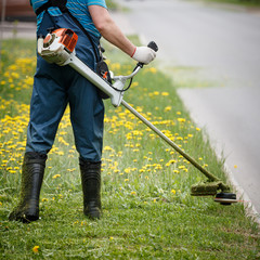 Closeup of a worker in special reflective clothing with a gasoline mower in hand. Man mows the...