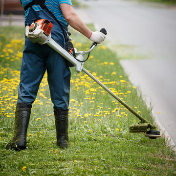 Closeup of a worker in special reflective clothing with a gasoline mower in hand. Man mows the grass with dandelions on the lawn with trimmer