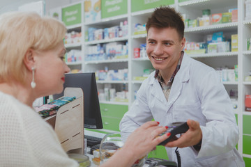 Senior womn paying for her purchase with a credit card at the drugstore. Cheerful male pharmacist...