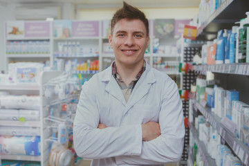 Happy handsome pharmacist standing with arms crossed at the drugstore. Friendly male chemist welcoming you in his pharmacy. Profession, occupation, medical service concept
