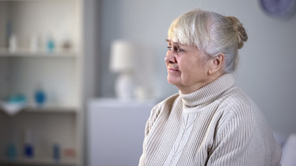 Fototapeta premium Depressed senior woman sitting alone in living room, abandoned by relatives