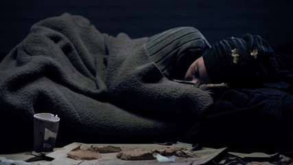 Homeless person covered by blanket sleeping on city street, poverty concept