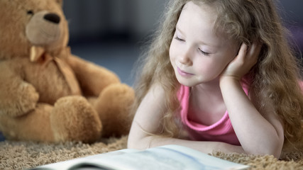 Fototapeta premium Concentrated cute girl reading book, lying on carpet, preschool education