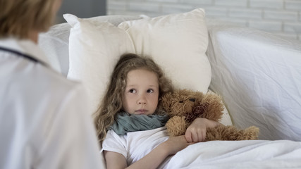 Sick girl looking at pediatrician in medical clinic, lying in bed, healthcare