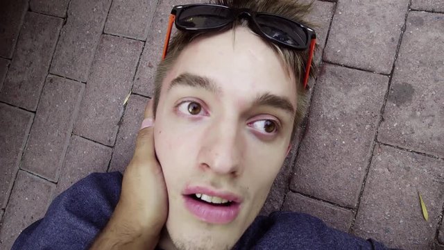 Handsome caucasian guy fainted guy with modern sunglasses is lying on concrete ground somewhere at street and being awaken with face slaps, he is opening his eyes in shock.