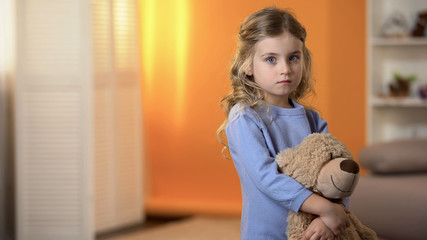 Adorable sad little girl hugging favorite teddy bear feeling lonely in orphanage