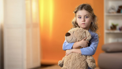 Abandoned upset curly girl holding favorite teddy bear feeling sad in orphanage