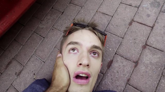 Attractive caucasian guy fainted guy with modern sunglasses is lying on concrete ground somewhere at street and being awaken with face slaps.