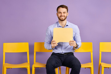 Happy young man sitting alone on yellow chair and holding up thought bubble and waiting for job interview