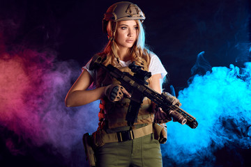 Fototapeta na wymiar Skilled blonde female soldier with rifle in hands standing in military outfit in smoky darkness. Woman, Military Service and firearm concept