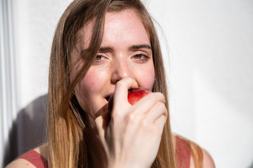 Young joyful attractive woman in casual summer dress sitting on balcony and eating tasty juicy red apple