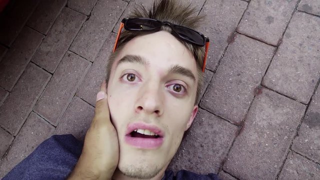 Cute caucasian fainted guy wearing modern sunglasses is lying on concrete ground in unconscious condition somewhere at street and being awaken with face slaps.