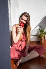 Young joyful attractive woman in casual summer dress sitting on balcony and eating tasty juicy red apple