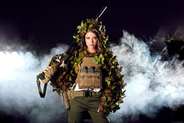 Camouflaged woman sniper in ghillie suit posing with rifle in foggy night. Special Weapons and...