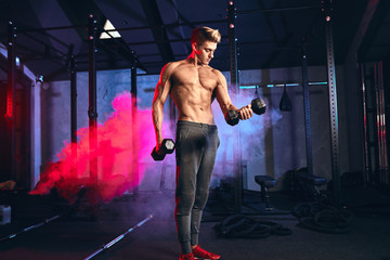 Caucasian man working out in gym doing exercises with dumbbells at triceps, strong male naked torso, highly defined muscles, six-packs slim trained body in dark gym with red spotlights