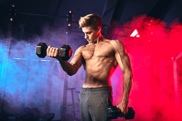 Shirtless white man in sweatpants starting exercise with dumbbell weight in dark gym with blue and red sportlight. Fitness motivation and muscle training concept.