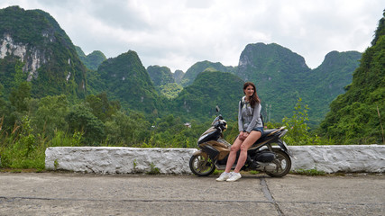 Young beautiful brunette woman sits and posing on the motorbike on the road with panoramic view to scenic landscape with green mountains. Cat Ba Island, Vietnam.