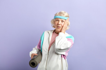 Surprised female pensioner holding yoga mat in hand, comes to gym with purple walls, being shocked...
