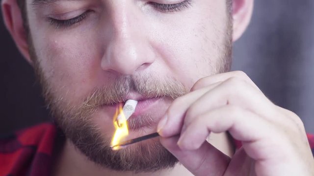 Portrait of mature nice-looking caucasian bearded guy with mustache who is slowly lighting and smoking small rolled cigarette and staring at camera with smile.