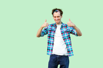 Portrait of satisfied young man in casual blue checkered shirt and headband standing, thumbs up and looking at camera with toothy smile. indoor studio shot, isolated on light green background.