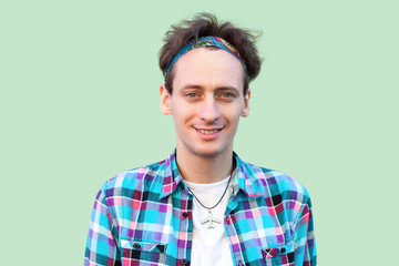 Closeup Portrait of happy successful young man in casual blue checkered shirt and headband standing and looking at camera with toothy smile. indoor studio shot, isolated on light green background.