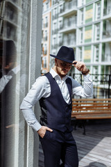 Portrait of handsome groom in dark-blue vest with hat on head in city center