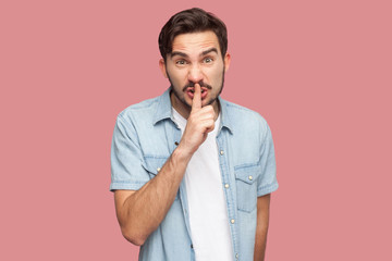 Portrait of serious or angry bearded young man in blue casual style shirt standing with silence sign gesture, finger on lips and looking at camera. indoor studio shot, isolated on pink background.