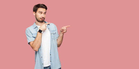 Portrait of attentive handsome bearded young man in blue casual style shirt standing, looking and pointing at background copyspace. indoor studio shot, isolated on pink background.