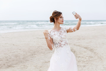 Fototapeta na wymiar Bride makes selfie on a smartphone on the sea beach. Wedding selfie on the background of the sea
