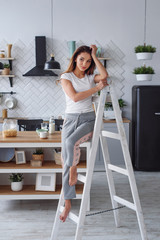 Positive beautiful young woman on a white wooden stepladder on the background of a stylish modern kitchen.