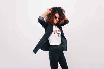 Slim girl with curly hairstyle laughing while dancing in studio. Charming tanned young lady in sunglasses and denim jacket having fun on white background.