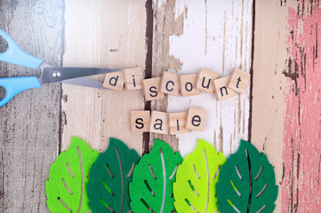 Concept image of price cut. Discount sale text being cut by blue color scissor over nice wooden texture background and green leaves prop